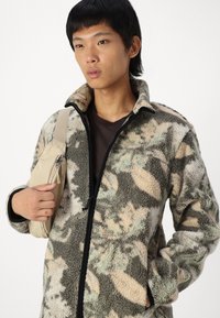 Fleece jacket in camouflage pattern with shades of green, beige, and brown. Features a zip front, high collar, and relaxed fit.