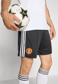 Juodos futbolo šortai su baltomis juostelėmis, ant kurių yra „Manchester United“ logotipas. Juoda ir balta futbolo kamuolys su žvaigždžių akcentais laikomas rankoje.