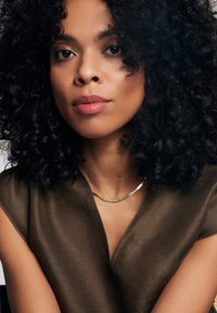 Brązowy, teksturowany top z dekoltem w kształcie litery V, zestawiony ze złotą, minimalistyczną naszyjnikiem. Kręcone czarne włosy otaczają twarz. Delikatny, neutralny makijaż.