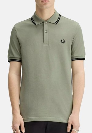 Uomo che indossa una maglietta polo a maniche corte color oliva chiaro con doppie strisce nere sul colletto e sui polsini, e un logo di corona d'alloro nero sul petto.
