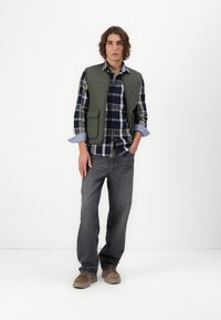 Pūkuveida zaļš vests virs šūnu krekla ar tumši zilu, baltu un zaļu toņu, saskaņots ar pelēkiem džinsiem un bēšiem sporta apaviem. Vienkāršs, funkcionāls dizains.