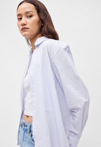 Πουκάμισο με μακριά μανίκια, με κουμπιά σε ανοιχτό μπλε χρώμα με λευκές ρίγες. Διαθέτει κλασικό γιακά και χαλαρή εφαρμογή, συνδυασμένο με λευκό tank top και τζιν παντελόνι.