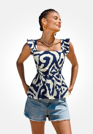 Vrouw met gevlochten haar draagt een marineblauw en wit topje zonder mouwen met gerimpelde bandjes, denim shorts, een schelpenketting en grote creolen.
