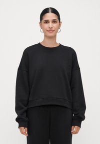 Mulher com cabelo escuro preso em um coque baixo, usando brincos de argola prateados, um moletom oversized preto e calças pretas combinando, de pé contra um fundo simples.