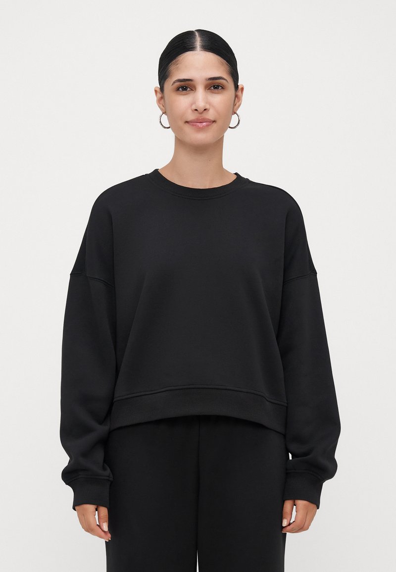 Mulher com cabelo escuro preso em um coque baixo, usando brincos de argola prateados, um moletom oversized preto e calças pretas combinando, de pé contra um fundo simples.