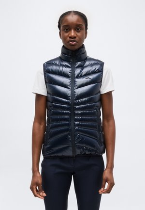 Γυναίκα που κοιτάζει ευθεία φορώντας ένα μπλε σκούρο αμάνικο καπιτονέ γιλέκο πάνω από ένα λευκό κοντομάνικο πουκάμισο και σκούρο παντελόνι, μπροστά σε απλό φόντο.