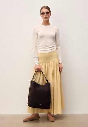 Naine, kandes ümmarguseid päikeseprille, valget pika varrukaga topi, kollast maksiseelikut, kootud baleriinakingi, hoides suurt musta käekotti tavalise seina taustal.