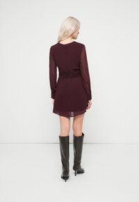 Βουργουνδί φόρεμα με μακριά διάφανα μανίκια και συγκεντρωμένη λεπτομέρεια στη μέση, συνδυασμένο με μαύρες μπότες μέχρι το γόνατο. Κοντό, ελαφρώς φουσκωτό στρίφωμα.