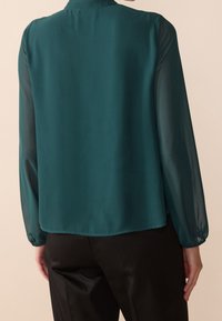 Blouse teal avec des manches longues et transparentes, de coupe légèrement ample, avec un col rond et une texture lisse et douce. Portée avec un pantalon noir.
