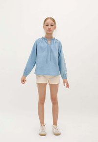 Jeune fille debout, portant une blouse à manches longues bleu clair avec des manches brodées, un short beige et des baskets blanches sur un fond uni.