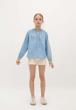 Jeune fille debout, portant une blouse à manches longues bleu clair avec des manches brodées, un short beige et des baskets blanches sur un fond uni.