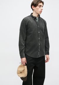 Sötétszürke kordbársony gombos ing terjedelmes gallérral és hosszú ujjal. A modellen bézs színű baseballsapka van. Fekete nadrágok egészítik ki a szettet.