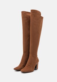 Bottes montantes en daim marron avec un bout pointu, un talon bloc et une texture lisse. Détail zippé sur le côté.