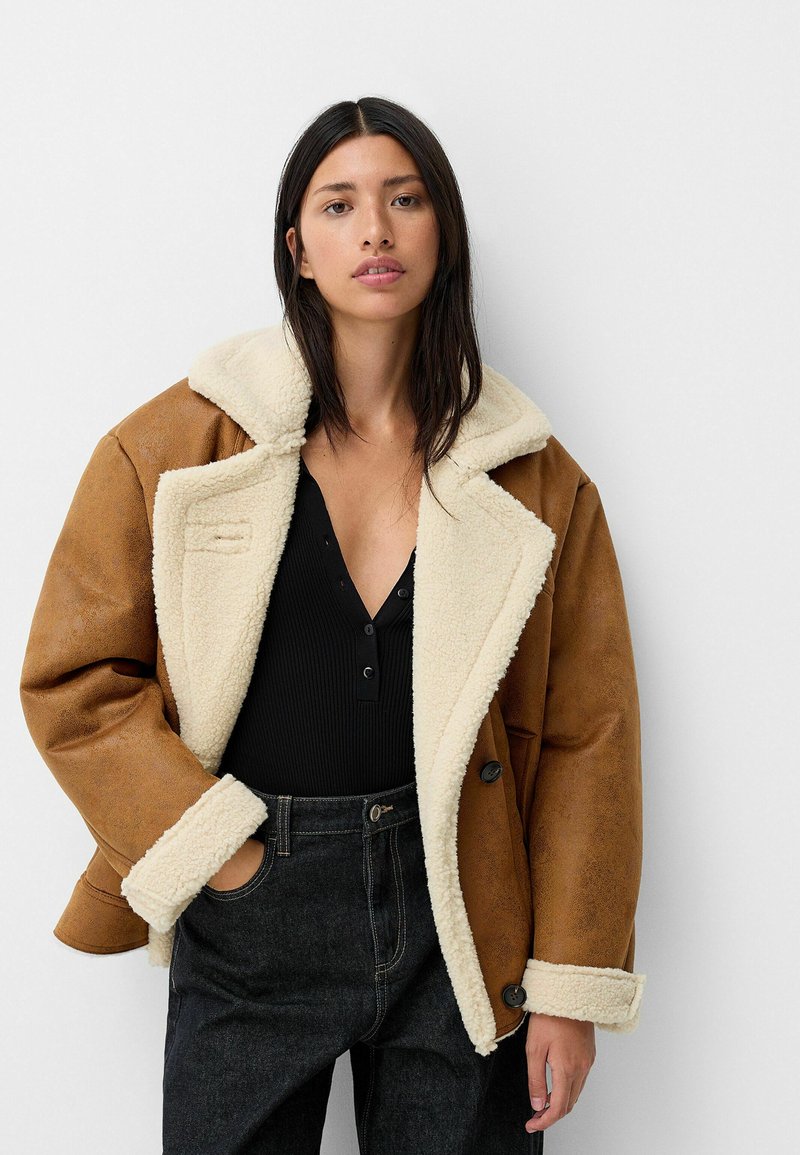Chaqueta de cuero sintético marrón con cuello de borreguillo crema, diseño de doble botonadura y botones negros, combinada con una blusa negra de canalé y jeans oscuros.