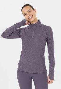 Ung kvinna som ler, iklädd en lila långärmad träningsblus med tumh�ål och matchande leggings mot en vit bakgrund.