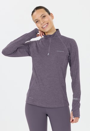 Jeune femme souriante, portant un haut de sport violet à manches longues avec des trous pour les pouces et un legging assorti sur fond blanc.