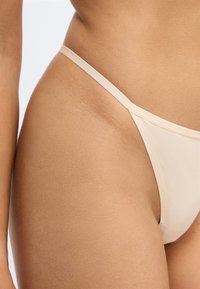 Bikini nude avec des fines bretelles, présentant une texture lisse et une couverture minimale. Le tissu semble léger et élastique.