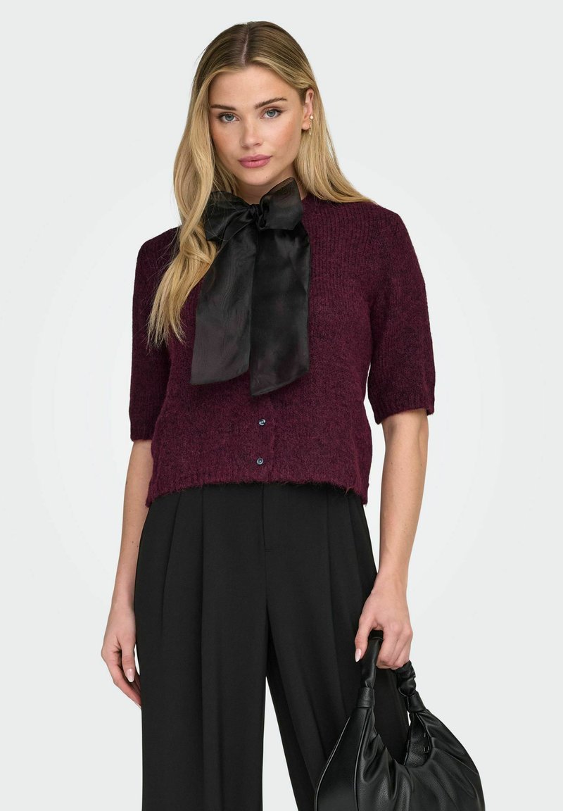 Maglione bordeaux a maniche corte con trama lavorata, caratterizzato da un fiocco in raso nero e chiusura frontale con tre bottoni.