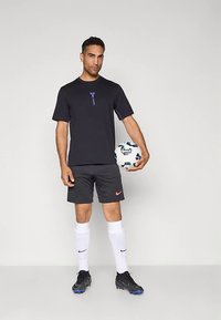 Mann i svart treningstøy holder en hvit og blå fotball, iført hvite Nike-sokker og svarte fotballsko, står foran en ensfarget bakgrunn.