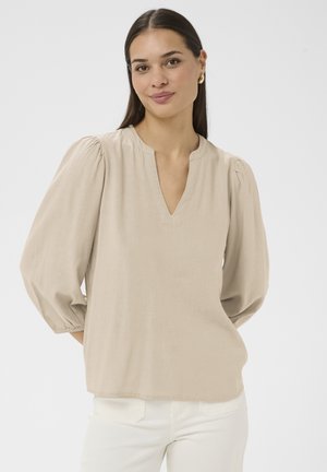 Femme aux longs cheveux foncés portant un chemisier beige à manches bouffantes avec col en V et un pantalon blanc, debout devant un fond clair uni.