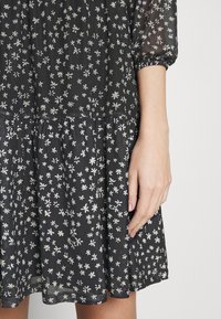 Svart klänning med vitt prästkrage-mönster, genomskinliga trekvarts puffärmar, med fokus på den nedre delen av torso och höger hand vilande vid sidan.