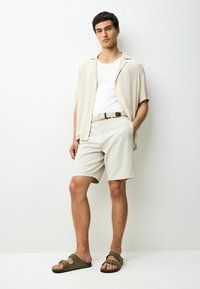 Pantaloni scurți dintr-un material bej deschis, asociați cu o cămașă crem lejeră și sandale maro. O curea împletită notabil accentuează talia.