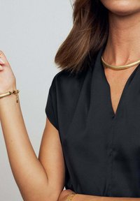 Sort kortærmet bluse med V-udskæring og glat tekstur, parret med guldsmykker, herunder en choker og et armbånd.