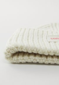 Kreemivärviline kootud tekk ribatekstuuriga, pehme villa-laadse materjaliga ja ühe nurga peal väikese logoplaadiga.