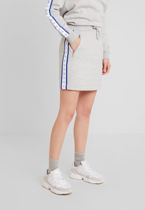 Kobieta ubrana w jasnoszarą spódnicę z bocznymi paskami i kieszenią na zamek, w zestawie z pasującą bluzą, szarymi skarpetkami i białymi sneakersami.