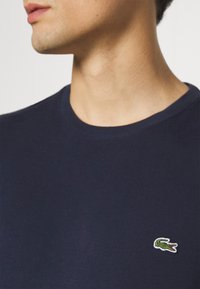 T-shirt à col rond bleu marine avec des manches courtes, présentant un petit logo de crocodile brodé sur la poitrine gauche. Texture de tissu lisse.