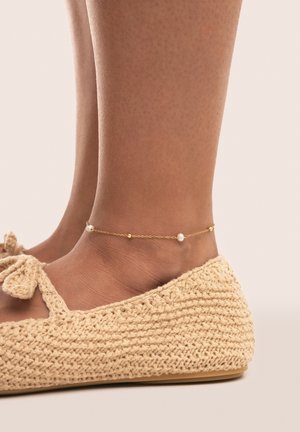 Zapatos planos de ganchillo en color beige con un lazo anudado presentan un tobillo adornado con una cadena dorada y una pequeña perla junto a cuentas doradas, descansando contra un tobillo suave.