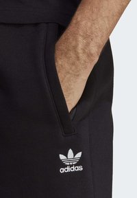 Černé bavlněné joggers s boční kapsou a bílým logem Adidas. Materiál má hladkou strukturu a volný střih v pase.