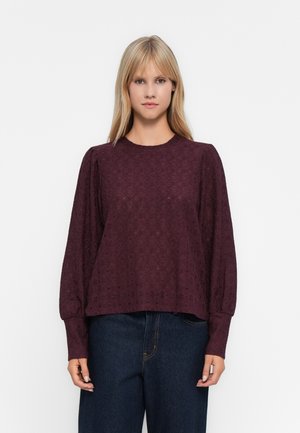 Kvinne iført en langermet, mørk lilla bluse i blomsterdelikat spiss med pufferte skuldre og mørkeblå høytlivsjeans mot en ensfarget bakgrunn.