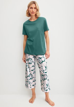 Frau steht barfuß, trägt ein türkisfarbenes Kurzarmoberteil und weit geschnittene, kurz geschnittene Hosen mit einem weißen und grünen Blumenmuster.