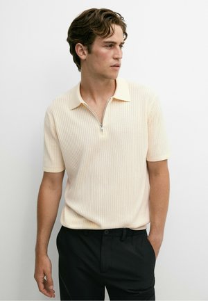 Junger Mann mit braunen Haaren, der ein kurzärmeliges beiges Polo-Shirt mit Reißverschluss und schwarze Hosen trägt, schaut seitlich vor weißem Hintergrund.
