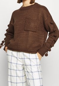 Pull en tricot marron avec une poche et des accents de pompon sur les manches. Porté avec un pantalon blanc à motif de grille bleue.