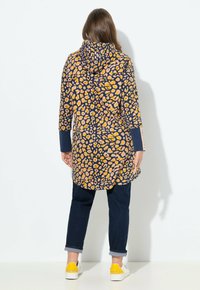 Veste à capuche avec un fond bleu marine arborant des taches de léopard jaunes et roses, à manches longues et avec des poches, associée à un jean foncé et des baskets.