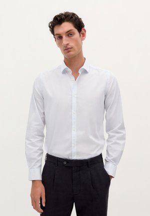Cămașă albă cu guler ascuțit și mâneci lungi, asortată cu pantaloni închiși la culoare. Material din bumbac neted, design minimalist.