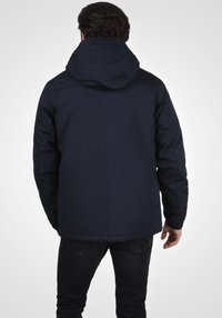 Mørkeblå isolert jakke med hette, glidelås, elastiske mansjetter og rett snitt. Enkel design, laget av værbestandig stoff.