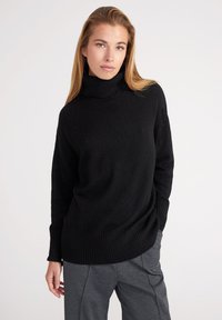 Pull à col roulé noir avec texture côtelée, manches longues et coupe décontractée, associé à un pantalon gris au design ajusté.