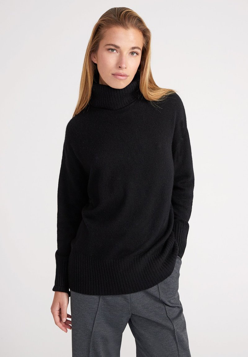 Pull à col roulé noir avec texture côtelée, manches longues et coupe décontractée, associé à un pantalon gris au design ajusté.