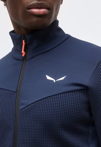 Άνδρας που φοράει ναυτικό μπλε υφασμάτινο μπουφάν με φερμουάρ, ψηλό γιακά και μικρό λευκό λογότυπο στο στήθος, ορατή κόκκινη τσίμπα φερμουάρ.