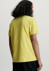 Žluté bavlněné tričko s krátkým rukávem a volným střihem, pohled ze zadu. Má jednobarevný vzhled a bezšvový design ramen.