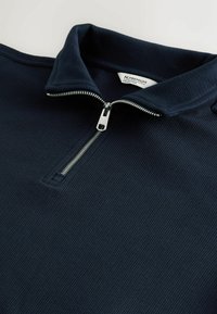 Tamsiai mėlynas, raštuotas audinys marškinėliai su sidabriniu pusiau užtrauktuku ir įlenktu apykakle, ant kurio matyti etiketė su užrašu „N. PREMIUM COLLECTION“.