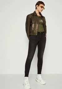 Žena v hnědém kabátu z umělé kůže, zeleném grafickém tričku, černých skinny džínsech, bílých mohutných teniskách a slunečních brýlích s oranžovým zabarvením, stojící před bílou zdí.