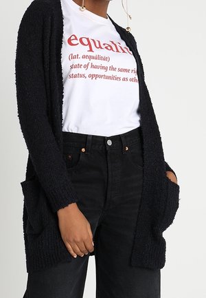 Personne portant un T-shirt blanc avec le texte « égalité » et sa définition en rouge, un cardigan noir et un jean noir taille haute, mains dans les poches.