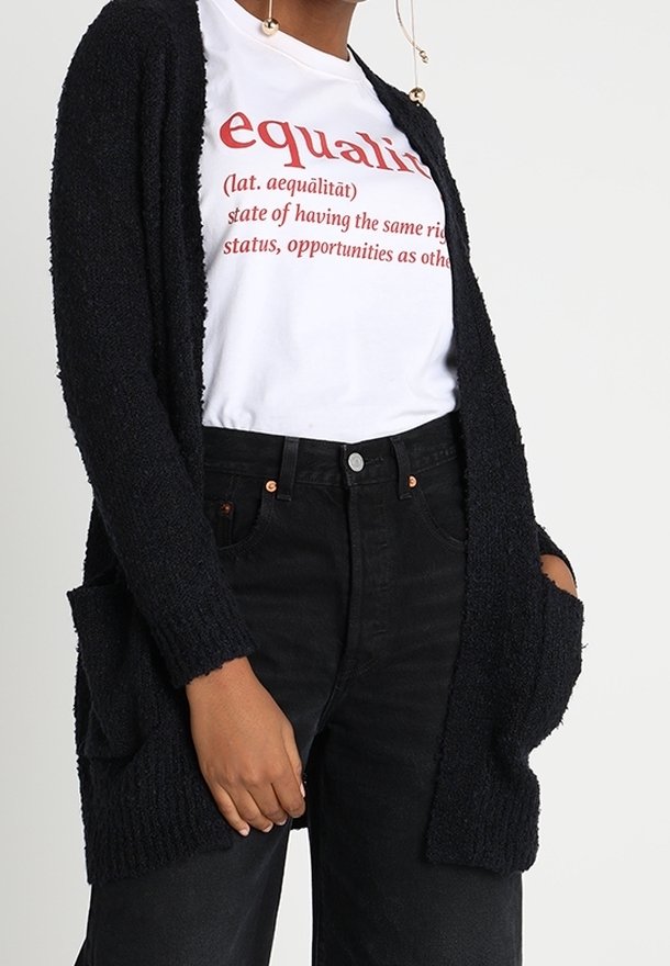 Personne portant un T-shirt blanc avec le texte « égalité » et sa définition en rouge, un cardigan noir et un jean noir taille haute, mains dans les poches.