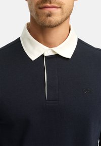 Marineblå polo skjorte med en kremfarget krage, knappelukking og strukturert stoff. Lite logo på brystet med en etablert dato.