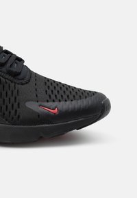 Svarta atletiska sko med en texturerad mesh-överdela med ventilationshål, gummisula och en röd swoosh-accent på sidan.