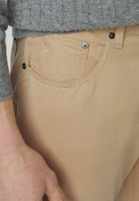 Tan bomuldsbukser med smal pasform, der har en frontlomme med broderet detalje, metalknaplukning og synlige stikninger.